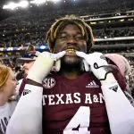 Ein Footballspieler, der ein Trikot von Texas A&M mit der Nummer 4 trägt und als Shemar Stewart identifiziert wurde, lächelt breit und präsentiert goldene Grillz. Mit einer verspielten Geste hält er seine Finger vor den Mund. Die Szene ist lebhaft, umgeben von Fans in einem Stadion voller Menschen unter hellen Lichtern in der Nacht. Diese Beschreibung wurde mit der FootballR KI automatisch generiert.