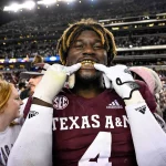 Ein Footballspieler, der ein Trikot von Texas A&M mit der Nummer 4 trägt und als Shemar Stewart identifiziert wurde, lächelt breit und präsentiert goldene Grillz. Mit einer verspielten Geste hält er seine Finger vor den Mund. Die Szene ist lebhaft, umgeben von Fans in einem Stadion voller Menschen unter hellen Lichtern in der Nacht. Diese Beschreibung wurde mit der FootballR KI automatisch generiert.