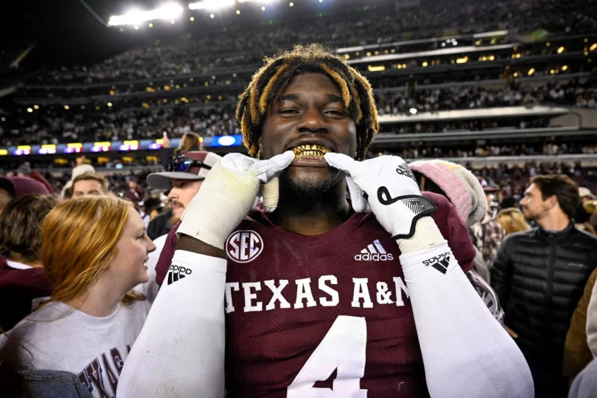 Ein Footballspieler, der ein Trikot von Texas A&M mit der Nummer 4 trägt und als Shemar Stewart identifiziert wurde, lächelt breit und präsentiert goldene Grillz. Mit einer verspielten Geste hält er seine Finger vor den Mund. Die Szene ist lebhaft, umgeben von Fans in einem Stadion voller Menschen unter hellen Lichtern in der Nacht. Diese Beschreibung wurde mit der FootballR KI automatisch generiert.