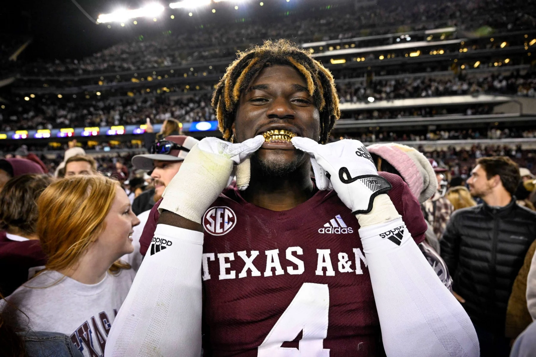 Ein Footballspieler, der ein Trikot von Texas A&M mit der Nummer 4 trägt und als Shemar Stewart identifiziert wurde, lächelt breit und präsentiert goldene Grillz. Mit einer verspielten Geste hält er seine Finger vor den Mund. Die Szene ist lebhaft, umgeben von Fans in einem Stadion voller Menschen unter hellen Lichtern in der Nacht. Diese Beschreibung wurde mit der FootballR KI automatisch generiert.