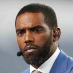 Randy Moss mit Bart und Schnurrbart, der an den legendären Stil von Randy Moss erinnert, trägt einen blauen Anzug und eine Krawatte. Er hat einen Ohrhörer mit angeschlossenem Mikrofon. Der Hintergrund ist verschwommen grau und sein Gesichtsausdruck ist ernst und konzentriert. Diese Beschreibung wurde mit der FootballR KI automatisch generiert.