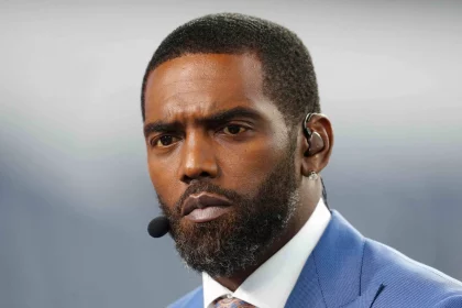 Randy Moss mit Bart und Schnurrbart, der an den legendären Stil von Randy Moss erinnert, trägt einen blauen Anzug und eine Krawatte. Er hat einen Ohrhörer mit angeschlossenem Mikrofon. Der Hintergrund ist verschwommen grau und sein Gesichtsausdruck ist ernst und konzentriert. Diese Beschreibung wurde mit der FootballR KI automatisch generiert.