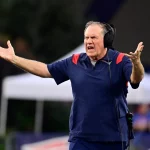 Ein Mann auf einem Footballfeld, der ein marineblaues Hemd mit roten Akzenten trägt, gestikuliert leidenschaftlich mit beiden erhobenen Armen wie Bill Belichick. Er trägt Kopfhörer und scheint seine Frustration auszudrücken oder Anweisungen zu geben. Im Hintergrund sind verschwommene Bäume und ein weißes Zelt zu sehen. Diese Beschreibung wurde mit der FootballR KI automatisch generiert.