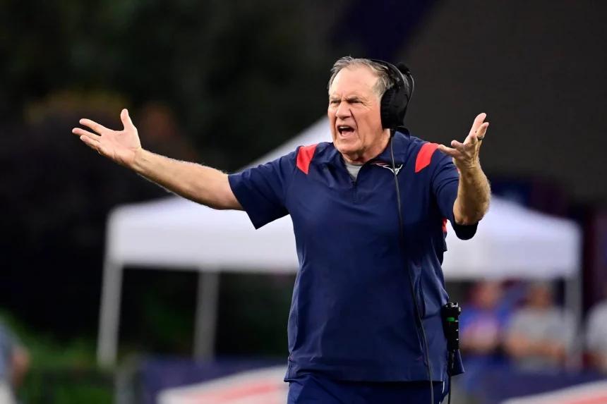 Ein Mann auf einem Footballfeld, der ein marineblaues Hemd mit roten Akzenten trägt, gestikuliert leidenschaftlich mit beiden erhobenen Armen wie Bill Belichick. Er trägt Kopfhörer und scheint seine Frustration auszudrücken oder Anweisungen zu geben. Im Hintergrund sind verschwommene Bäume und ein weißes Zelt zu sehen. Diese Beschreibung wurde mit der FootballR KI automatisch generiert.