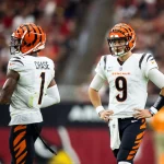 Zwei Footballspieler der Cincinnati Bengals stehen auf dem Feld. Der Spieler links mit der Trikotnummer 1 - Ja'Marr Chase - blickt in Richtung Endzone. Der Spieler rechts ist Joe Burrow mit der Trikotnummer 9, der mit den Händen auf den Hüften steht. Beide tragen weiße Trikots, schwarz-orange gestreifte Helme und orangefarbene Hosen. Diese Beschreibung wurde mit der FootballR KI automatisch generiert.