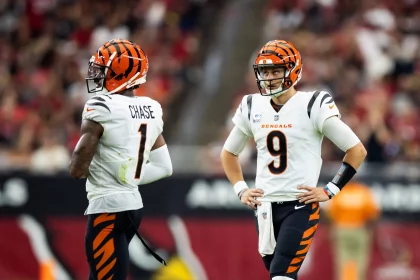 Zwei Footballspieler der Cincinnati Bengals stehen auf dem Feld. Der Spieler links mit der Trikotnummer 1 - Ja'Marr Chase - blickt in Richtung Endzone. Der Spieler rechts ist Joe Burrow mit der Trikotnummer 9, der mit den Händen auf den Hüften steht. Beide tragen weiße Trikots, schwarz-orange gestreifte Helme und orangefarbene Hosen. Diese Beschreibung wurde mit der FootballR KI automatisch generiert.