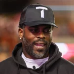 Neo Norfolk State Coach Michael Vick mit schwarzer Baseballkappe mit weißem „T“-Logo und schwarzem Kapuzenpullover lächelt und unterhält sich. Die verschwommenen, hellen Lichter deuten auf eine lebhafte Umgebung hin. Mit Bart und Ohrsteckern könnte man sie leicht mit einer NFL-Legende verwechseln, die die Nacht genießt. Diese Beschreibung wurde mit der FootballR KI automatisch generiert.