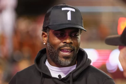 Neo Norfolk State Coach Michael Vick mit schwarzer Baseballkappe mit weißem „T“-Logo und schwarzem Kapuzenpullover lächelt und unterhält sich. Die verschwommenen, hellen Lichter deuten auf eine lebhafte Umgebung hin. Mit Bart und Ohrsteckern könnte man sie leicht mit einer NFL-Legende verwechseln, die die Nacht genießt. Diese Beschreibung wurde mit der FootballR KI automatisch generiert.