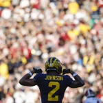 Ein Footballspieler, Will Johnson, trägt eine dunkelblaue und gelbe Uniform mit der Nummer 2 und steht vor einer verschwommenen jubelnden Menge. Er zeigt stolz auf seinen Namen. Das markante gelbe Muster auf blauem Grund verleiht dem Helm Flair und fängt die Spannung des Spiels perfekt ein. Diese Beschreibung wurde mit der FootballR KI automatisch generiert.