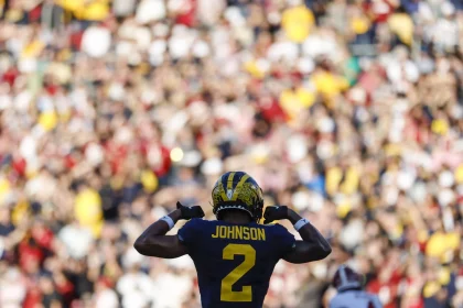 Ein Footballspieler, Will Johnson, trägt eine dunkelblaue und gelbe Uniform mit der Nummer 2 und steht vor einer verschwommenen jubelnden Menge. Er zeigt stolz auf seinen Namen. Das markante gelbe Muster auf blauem Grund verleiht dem Helm Flair und fängt die Spannung des Spiels perfekt ein. Diese Beschreibung wurde mit der FootballR KI automatisch generiert.