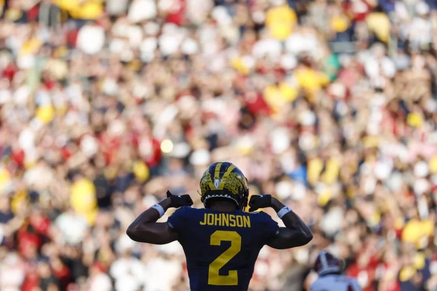 Ein Footballspieler, Will Johnson, trägt eine dunkelblaue und gelbe Uniform mit der Nummer 2 und steht vor einer verschwommenen jubelnden Menge. Er zeigt stolz auf seinen Namen. Das markante gelbe Muster auf blauem Grund verleiht dem Helm Flair und fängt die Spannung des Spiels perfekt ein. Diese Beschreibung wurde mit der FootballR KI automatisch generiert.