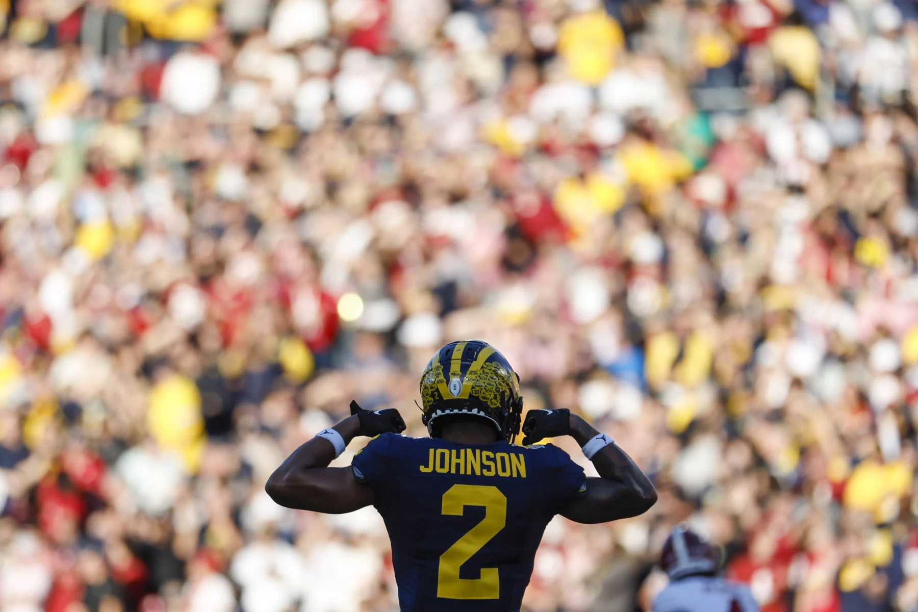 Ein Footballspieler, Will Johnson, trägt eine dunkelblaue und gelbe Uniform mit der Nummer 2 und steht vor einer verschwommenen jubelnden Menge. Er zeigt stolz auf seinen Namen. Das markante gelbe Muster auf blauem Grund verleiht dem Helm Flair und fängt die Spannung des Spiels perfekt ein. Diese Beschreibung wurde mit der FootballR KI automatisch generiert.