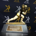 Travis Hunter oder Ashton Jeanty? Wer gewinnt? Das Bild zeigt die Heisman Trophy 2024 mit ihrer ikonischen Bronzestatue eines Footballspielers in dynamischer Laufpose mit ausgestrecktem Arm. Sie ruht auf einem Holzsockel mit einer Plakette. Dahinter ist ein schwarzer Vorhang mit goldenen Umrissen derselben berühmten Statue geschmückt. Diese Beschreibung wurde mit der FootballR KI automatisch generiert.