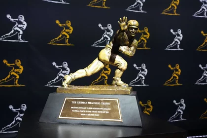 Travis Hunter oder Ashton Jeanty? Wer gewinnt? Das Bild zeigt die Heisman Trophy 2024 mit ihrer ikonischen Bronzestatue eines Footballspielers in dynamischer Laufpose mit ausgestrecktem Arm. Sie ruht auf einem Holzsockel mit einer Plakette. Dahinter ist ein schwarzer Vorhang mit goldenen Umrissen derselben berühmten Statue geschmückt. Diese Beschreibung wurde mit der FootballR KI automatisch generiert.