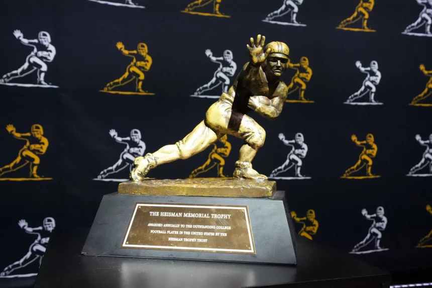Travis Hunter oder Ashton Jeanty? Wer gewinnt? Das Bild zeigt die Heisman Trophy 2024 mit ihrer ikonischen Bronzestatue eines Footballspielers in dynamischer Laufpose mit ausgestrecktem Arm. Sie ruht auf einem Holzsockel mit einer Plakette. Dahinter ist ein schwarzer Vorhang mit goldenen Umrissen derselben berühmten Statue geschmückt. Diese Beschreibung wurde mit der FootballR KI automatisch generiert.
