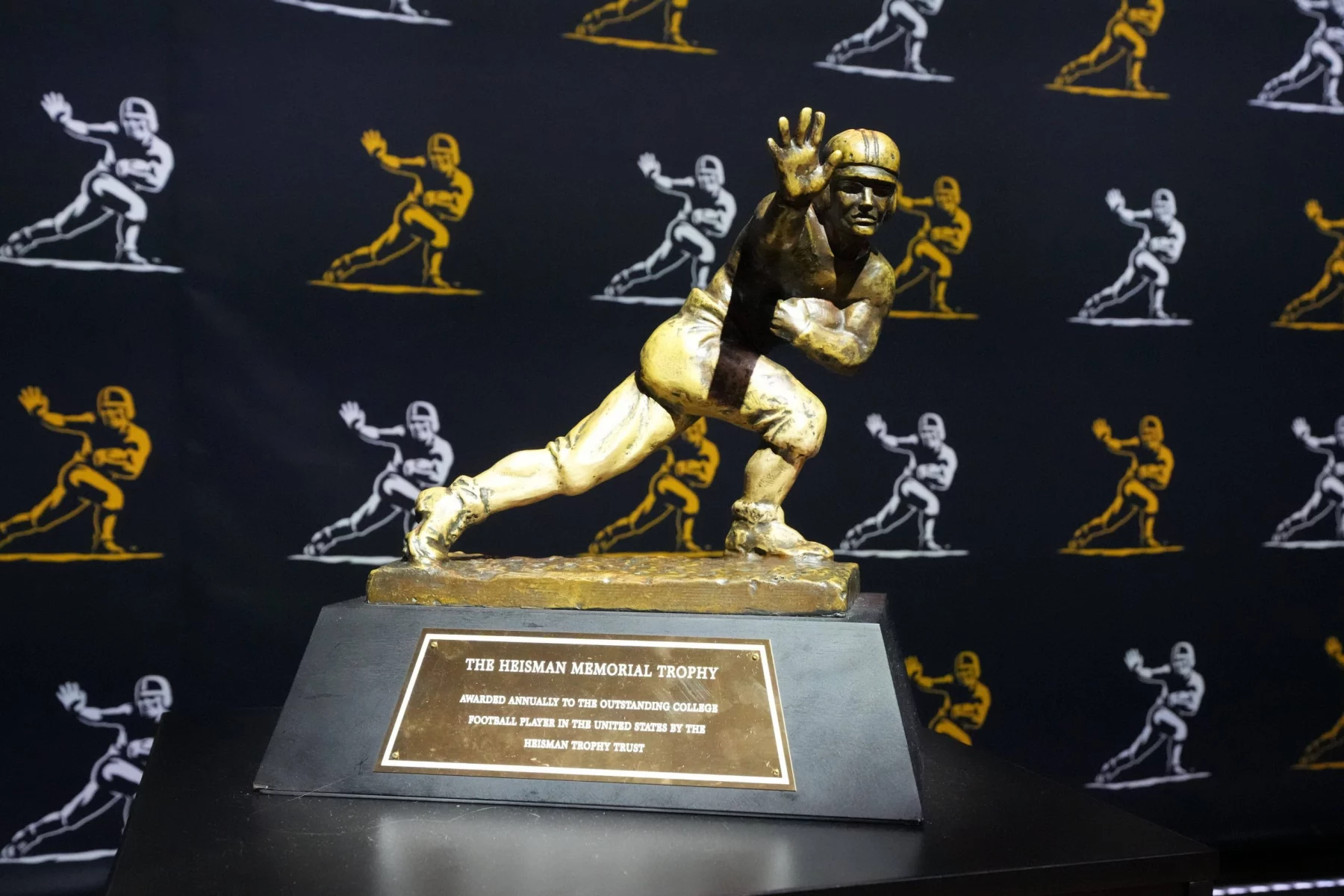 Travis Hunter oder Ashton Jeanty? Wer gewinnt? Das Bild zeigt die Heisman Trophy 2024 mit ihrer ikonischen Bronzestatue eines Footballspielers in dynamischer Laufpose mit ausgestrecktem Arm. Sie ruht auf einem Holzsockel mit einer Plakette. Dahinter ist ein schwarzer Vorhang mit goldenen Umrissen derselben berühmten Statue geschmückt. Diese Beschreibung wurde mit der FootballR KI automatisch generiert.