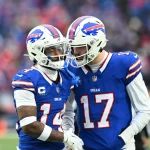 Stefon Diggs und Josh Allen in blauen Buffalo Bills-Trikots verbringen einen Moment auf dem Spielfeld. Einer trägt die Nummer 14, Stefon Diggs, und der andere, NFL MVP Kandidat Josh Allen, trägt die Nummer 17. Beide tragen weiße Helme mit einem rot-blauen Logo und stehen dicht beieinander in einem Stadion voller verschwommener Zuschauer. Diese Beschreibung wurde mit der FootballR KI automatisch generiert.