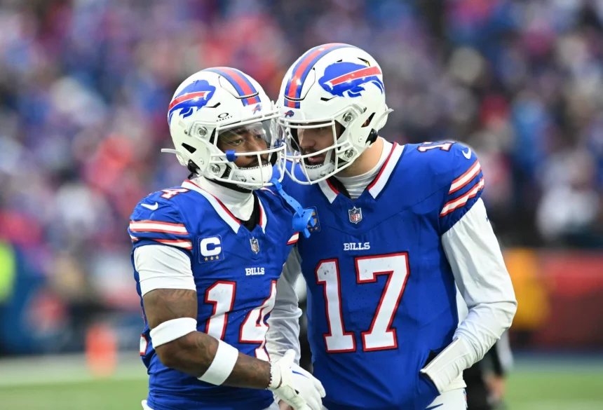 Stefon Diggs und Josh Allen in blauen Buffalo Bills-Trikots verbringen einen Moment auf dem Spielfeld. Einer trägt die Nummer 14, Stefon Diggs, und der andere, NFL MVP Kandidat Josh Allen, trägt die Nummer 17. Beide tragen weiße Helme mit einem rot-blauen Logo und stehen dicht beieinander in einem Stadion voller verschwommener Zuschauer. Diese Beschreibung wurde mit der FootballR KI automatisch generiert.