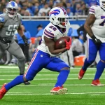 DAZN NFL Programm Woche 15: Ein Footballspieler im weißen Trikot mit der Nummer 26 sprintet mit dem Ball über das Feld. Sein Helm glänzt und seine blauen Hosen passen zu seinen schnellen Schritten. Gegnerische Spieler in grauen Uniformen jagen ihn, während ein mit Zuschauern vollgestopftes Stadion summt und an die Atmosphäre eines NFL-Livespiels auf DAZN erinnert. Diese Beschreibung wurde mit der FootballR KI automatisch generiert.