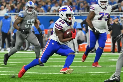 DAZN NFL Programm Woche 15: Ein Footballspieler im weißen Trikot mit der Nummer 26 sprintet mit dem Ball über das Feld. Sein Helm glänzt und seine blauen Hosen passen zu seinen schnellen Schritten. Gegnerische Spieler in grauen Uniformen jagen ihn, während ein mit Zuschauern vollgestopftes Stadion summt und an die Atmosphäre eines NFL-Livespiels auf DAZN erinnert. Diese Beschreibung wurde mit der FootballR KI automatisch generiert.