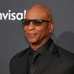 Eric Dickerson in schwarzem Anzug, schwarzem Hemd und schwarzer Krawatte lächelt selbstbewusst. Sie trägt eine dunkle Sonnenbrille und steht vor einem dunklen Hintergrund, auf dem ein Teil des Wortes „invisible“ zu sehen ist und auf der linken Seite ein Teil des Logos, was an Saquon Barkley's eleganten Stil erinnert. Diese Beschreibung wurde mit der FootballR KI automatisch generiert.