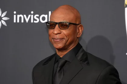 Eric Dickerson in schwarzem Anzug, schwarzem Hemd und schwarzer Krawatte lächelt selbstbewusst. Sie trägt eine dunkle Sonnenbrille und steht vor einem dunklen Hintergrund, auf dem ein Teil des Wortes „invisible“ zu sehen ist und auf der linken Seite ein Teil des Logos, was an Saquon Barkley's eleganten Stil erinnert. Diese Beschreibung wurde mit der FootballR KI automatisch generiert.