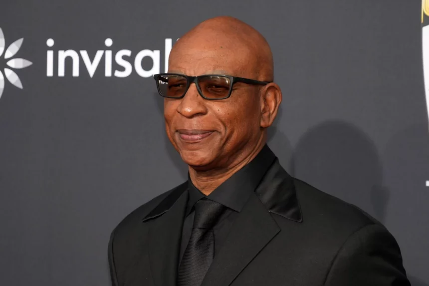 Eric Dickerson in schwarzem Anzug, schwarzem Hemd und schwarzer Krawatte lächelt selbstbewusst. Sie trägt eine dunkle Sonnenbrille und steht vor einem dunklen Hintergrund, auf dem ein Teil des Wortes „invisible“ zu sehen ist und auf der linken Seite ein Teil des Logos, was an Saquon Barkley's eleganten Stil erinnert. Diese Beschreibung wurde mit der FootballR KI automatisch generiert.