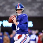 Eli Manning in blau-roter Uniform, die an Eli Mannings ikonischen Stil erinnert, hält einen Football in gespannter Position, bereit zum Werfen. Er ist auf dem Feld, im Hintergrund sind Zuschauer und Spieler verschwommen zu sehen. Die Trikotnummer des Spielers ist 12. Es ist eindeutig Spielzeit oder eine Trainingseinheit. Diese Beschreibung wurde mit der FootballR KI automatisch generiert.