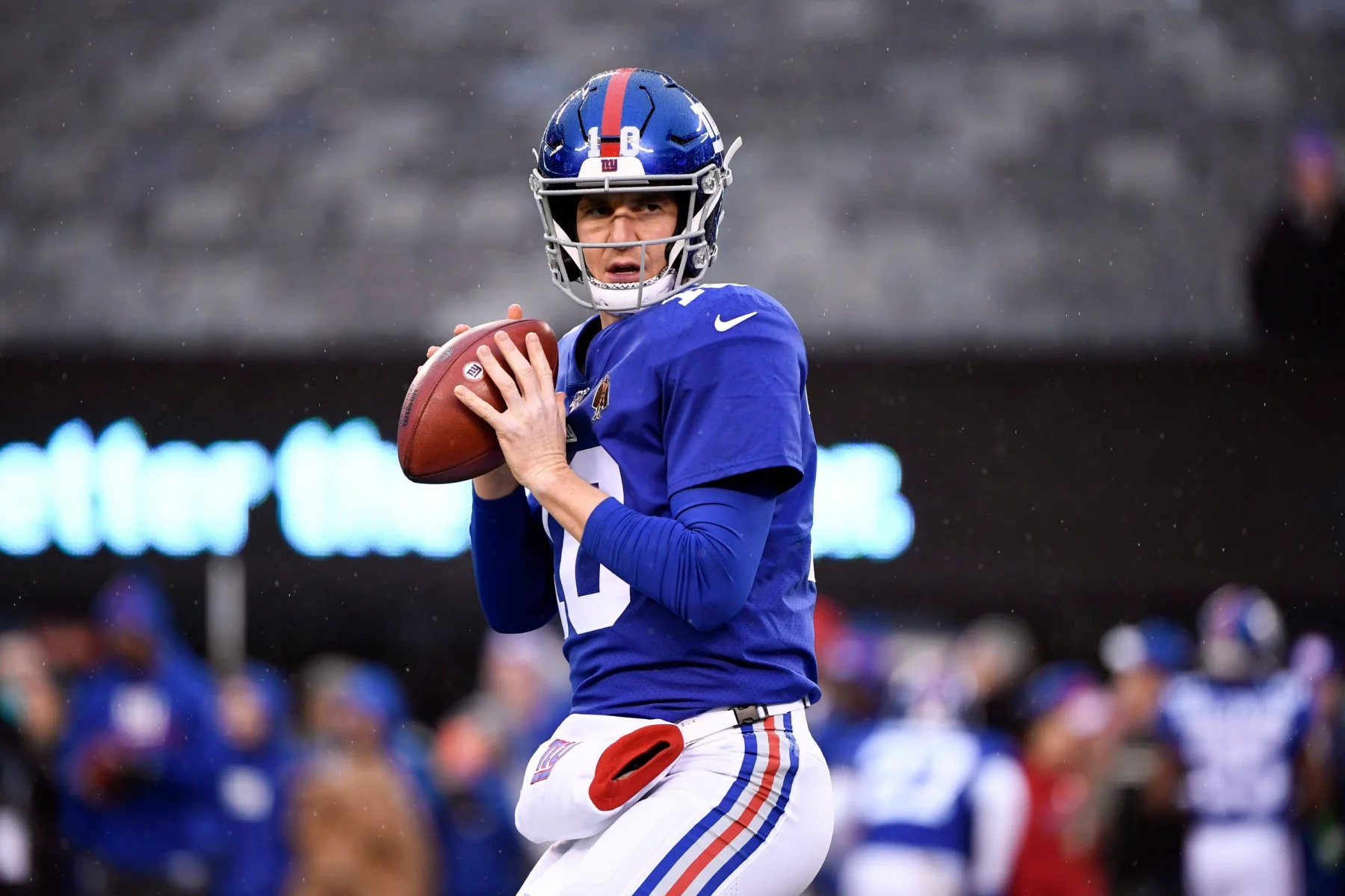 Eli Manning in blau-roter Uniform, die an Eli Mannings ikonischen Stil erinnert, hält einen Football in gespannter Position, bereit zum Werfen. Er ist auf dem Feld, im Hintergrund sind Zuschauer und Spieler verschwommen zu sehen. Die Trikotnummer des Spielers ist 12. Es ist eindeutig Spielzeit oder eine Trainingseinheit. Diese Beschreibung wurde mit der FootballR KI automatisch generiert.