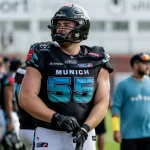 Ein Footballspieler, Leander Wiegand, trägt einen schwarzen Helm und ein Trikot mit der Nummer 55 und der Aufschrift „MÜNCHEN“ und steht auf einem Feld und richtet seine Handschuhe. Im Hintergrund dieser hellen Freiluftsportanlage sind weitere Spieler und Mitarbeiter in ähnlichen Uniformen zu sehen. Diese Beschreibung wurde mit der FootballR KI automatisch generiert.