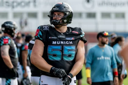 Ein Footballspieler, Leander Wiegand, trägt einen schwarzen Helm und ein Trikot mit der Nummer 55 und der Aufschrift „MÜNCHEN“ und steht auf einem Feld und richtet seine Handschuhe. Im Hintergrund dieser hellen Freiluftsportanlage sind weitere Spieler und Mitarbeiter in ähnlichen Uniformen zu sehen. Diese Beschreibung wurde mit der FootballR KI automatisch generiert.