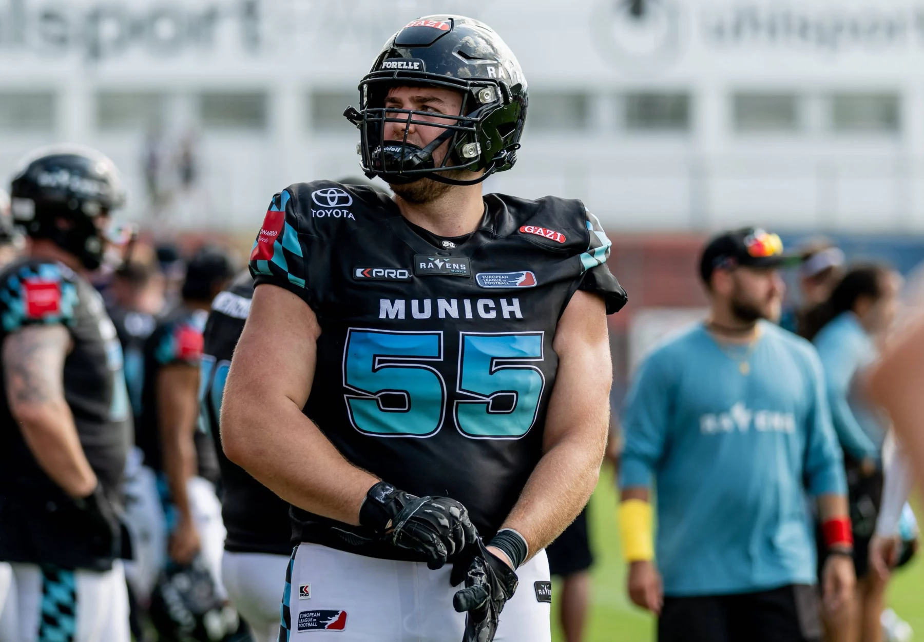 Ein Footballspieler, Leander Wiegand, trägt einen schwarzen Helm und ein Trikot mit der Nummer 55 und der Aufschrift „MÜNCHEN“ und steht auf einem Feld und richtet seine Handschuhe. Im Hintergrund dieser hellen Freiluftsportanlage sind weitere Spieler und Mitarbeiter in ähnlichen Uniformen zu sehen. Diese Beschreibung wurde mit der FootballR KI automatisch generiert.