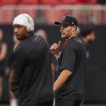 Zwei Männer in schwarzer Sportkleidung - Kirk Cousins und Michael Penix Jr. - stehen am Feld. Einer trägt eine Kappe und gestikuliert mit dem Daumen nach hinten. Der andere, der Kopfhörer und eine Kappe trägt, blickt nach vorne, ist aber verschwommen. Ein Sportplatz und mehrere Personen im Hintergrund sind leicht unscharf. Diese Beschreibung wurde mit der FootballR KI automatisch generiert.