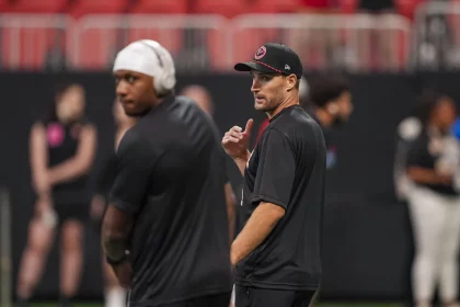 Zwei Männer in schwarzer Sportkleidung - Kirk Cousins und Michael Penix Jr. - stehen am Feld. Einer trägt eine Kappe und gestikuliert mit dem Daumen nach hinten. Der andere, der Kopfhörer und eine Kappe trägt, blickt nach vorne, ist aber verschwommen. Ein Sportplatz und mehrere Personen im Hintergrund sind leicht unscharf. Diese Beschreibung wurde mit der FootballR KI automatisch generiert.