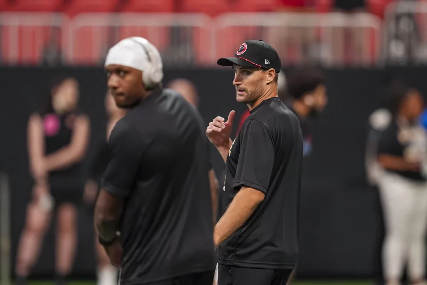 Zwei Männer in schwarzer Sportkleidung - Kirk Cousins und Michael Penix Jr. - stehen am Feld. Einer trägt eine Kappe und gestikuliert mit dem Daumen nach hinten. Der andere, der Kopfhörer und eine Kappe trägt, blickt nach vorne, ist aber verschwommen. Ein Sportplatz und mehrere Personen im Hintergrund sind leicht unscharf. Diese Beschreibung wurde mit der FootballR KI automatisch generiert.