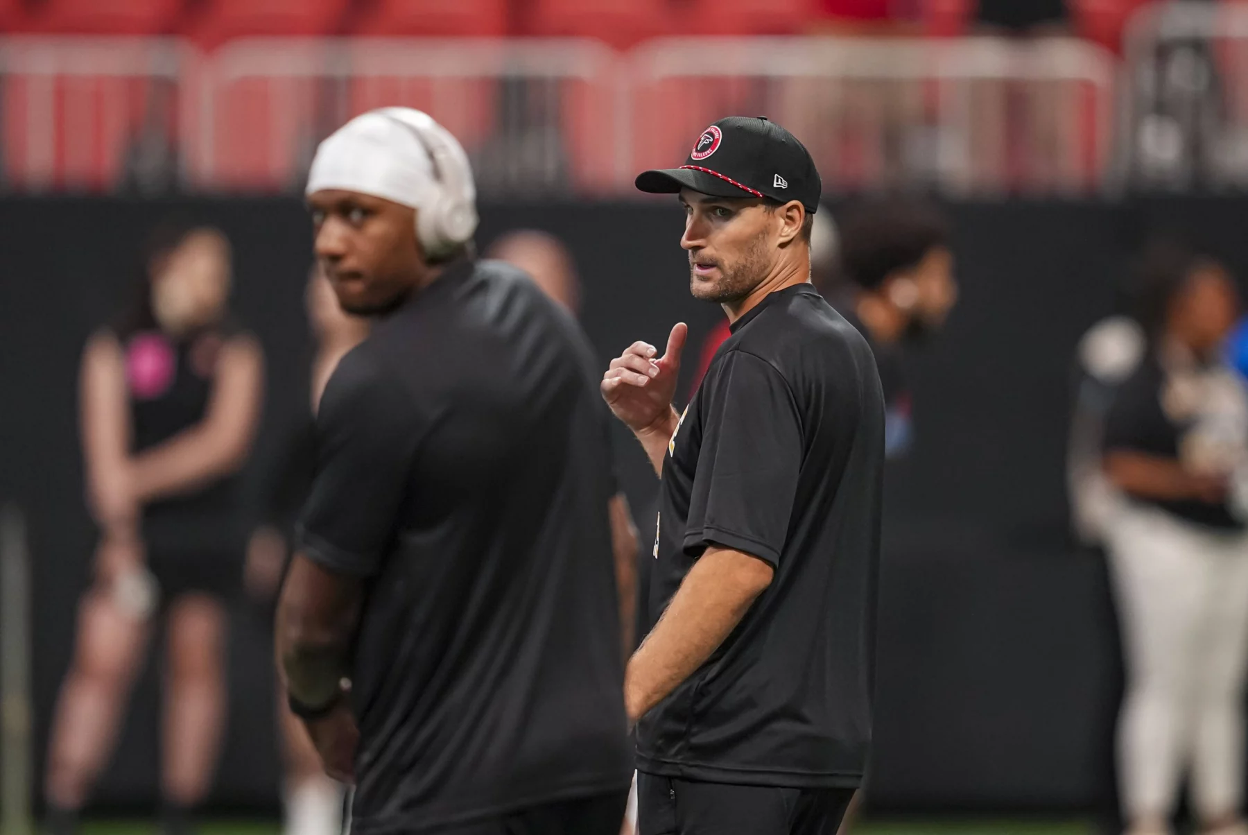 Zwei Männer in schwarzer Sportkleidung - Kirk Cousins und Michael Penix Jr. - stehen am Feld. Einer trägt eine Kappe und gestikuliert mit dem Daumen nach hinten. Der andere, der Kopfhörer und eine Kappe trägt, blickt nach vorne, ist aber verschwommen. Ein Sportplatz und mehrere Personen im Hintergrund sind leicht unscharf. Diese Beschreibung wurde mit der FootballR KI automatisch generiert.