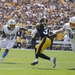 Es läuft eine Footballszene ab, in der J.K. Dobbins, Nummer 27 in Weiß, das Spielfeld hinuntersprintet, während ein Spieler in schwarz-gelber Uniform, Nummer 31, zum Tackling herankommt. Ein anderer Spieler in Weiß, Nummer 81, rennt los, um zu blocken. Das Stadion summt vor Vorfreude der dicht gedrängten Menge von Fans. Diese Beschreibung wurde mit der FootballR KI automatisch generiert.