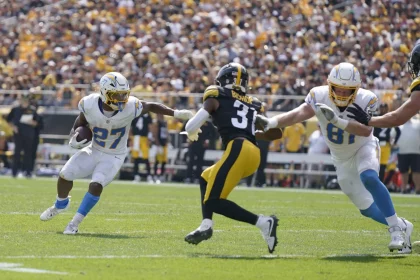 Es läuft eine Footballszene ab, in der J.K. Dobbins, Nummer 27 in Weiß, das Spielfeld hinuntersprintet, während ein Spieler in schwarz-gelber Uniform, Nummer 31, zum Tackling herankommt. Ein anderer Spieler in Weiß, Nummer 81, rennt los, um zu blocken. Das Stadion summt vor Vorfreude der dicht gedrängten Menge von Fans. Diese Beschreibung wurde mit der FootballR KI automatisch generiert.