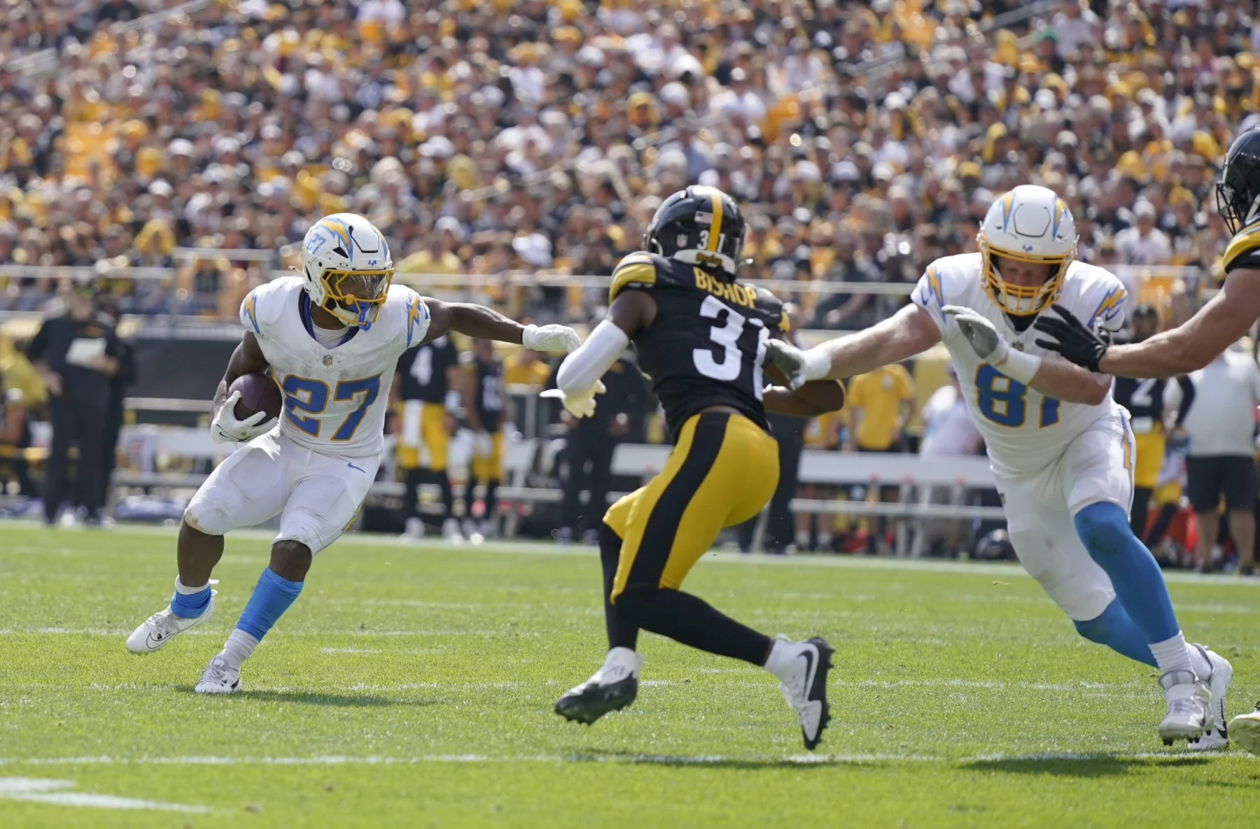 Es läuft eine Footballszene ab, in der J.K. Dobbins, Nummer 27 in Weiß, das Spielfeld hinuntersprintet, während ein Spieler in schwarz-gelber Uniform, Nummer 31, zum Tackling herankommt. Ein anderer Spieler in Weiß, Nummer 81, rennt los, um zu blocken. Das Stadion summt vor Vorfreude der dicht gedrängten Menge von Fans. Diese Beschreibung wurde mit der FootballR KI automatisch generiert.