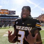 Ein Footballspieler, Luther Burden III, trägt ein schwarz-goldenes „Mizzou“-Trikot mit der Nummer 3 und steht stolz auf dem Spielfeld. Lächelnd und mit beiden Händen gestikulierend genießt er den Moment, während auf der Anzeigetafel unter einem klaren Himmel vor einer begeisterten Menge „Mizzou Bowl Eligible!“ steht. Diese Beschreibung wurde mit der FootballR KI automatisch generiert.