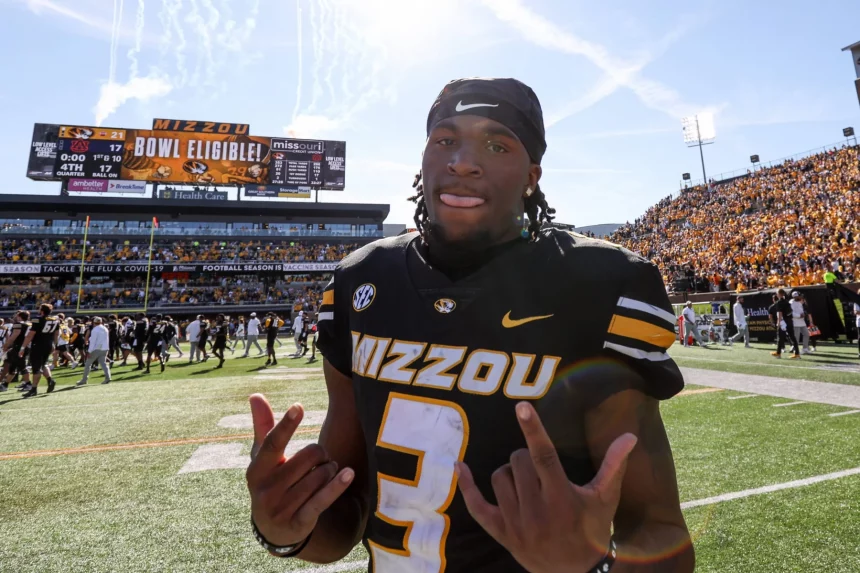 Ein Footballspieler, Luther Burden III, trägt ein schwarz-goldenes „Mizzou“-Trikot mit der Nummer 3 und steht stolz auf dem Spielfeld. Lächelnd und mit beiden Händen gestikulierend genießt er den Moment, während auf der Anzeigetafel unter einem klaren Himmel vor einer begeisterten Menge „Mizzou Bowl Eligible!“ steht. Diese Beschreibung wurde mit der FootballR KI automatisch generiert.