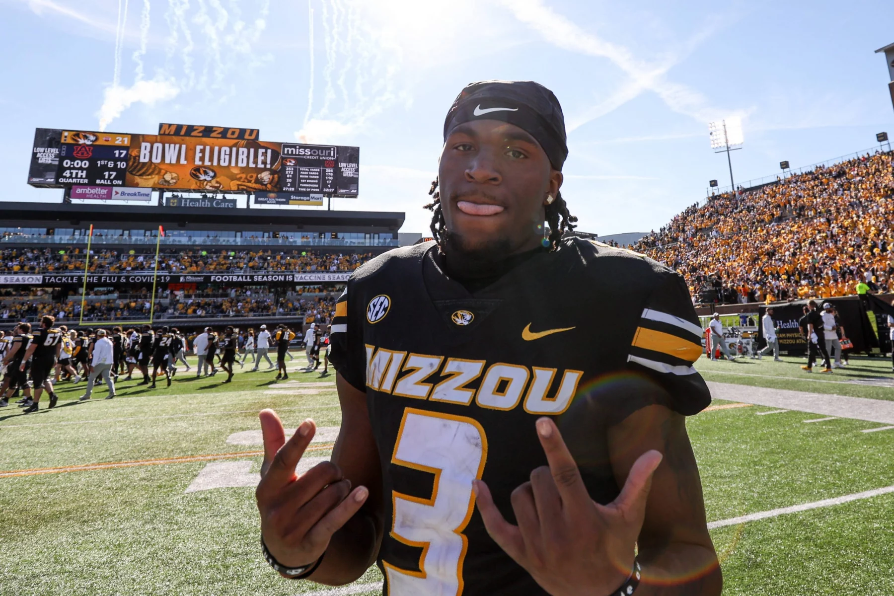 Ein Footballspieler, Luther Burden III, trägt ein schwarz-goldenes „Mizzou“-Trikot mit der Nummer 3 und steht stolz auf dem Spielfeld. Lächelnd und mit beiden Händen gestikulierend genießt er den Moment, während auf der Anzeigetafel unter einem klaren Himmel vor einer begeisterten Menge „Mizzou Bowl Eligible!“ steht. Diese Beschreibung wurde mit der FootballR KI automatisch generiert.