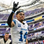 NFL Spielplan Woche 18: Amon-Ra St. Brown in der Uniform des Detroit-Teams mit der Nummer 14 und einem Kapitänsabzeichen hebt während des NFL-Spielplans Lions vs. Vikings seine Hand auf dem Spielfeld. Er trägt ein schwarzes Stirnband und Handschuhe, während im verschwommenen Hintergrund des Stadions eine weitere Person in einer grauen Weste in der Nähe steht. Diese Beschreibung wurde mit der FootballR KI automatisch generiert.