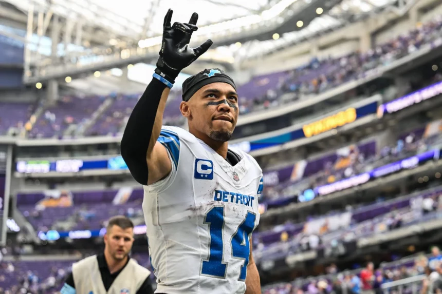 NFL Spielplan Woche 18: Amon-Ra St. Brown in der Uniform des Detroit-Teams mit der Nummer 14 und einem Kapitänsabzeichen hebt während des NFL-Spielplans Lions vs. Vikings seine Hand auf dem Spielfeld. Er trägt ein schwarzes Stirnband und Handschuhe, während im verschwommenen Hintergrund des Stadions eine weitere Person in einer grauen Weste in der Nähe steht. Diese Beschreibung wurde mit der FootballR KI automatisch generiert.