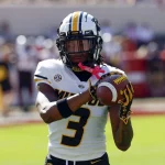 Luther Burden III trägt eine schwarz-weiße Uniform mit der Nummer 3 und demonstriert explosives Potenzial, als er einen Football fängt. Seine Handschuhe weisen rosa Akzente auf. Das verschwommene Spielfeld und die Stadionsitze deuten an diesem sonnigen Tag das Rot im Hintergrund an. Diese Beschreibung wurde mit der FootballR KI automatisch generiert.