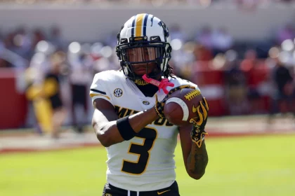 Luther Burden III trägt eine schwarz-weiße Uniform mit der Nummer 3 und demonstriert explosives Potenzial, als er einen Football fängt. Seine Handschuhe weisen rosa Akzente auf. Das verschwommene Spielfeld und die Stadionsitze deuten an diesem sonnigen Tag das Rot im Hintergrund an. Diese Beschreibung wurde mit der FootballR KI automatisch generiert.