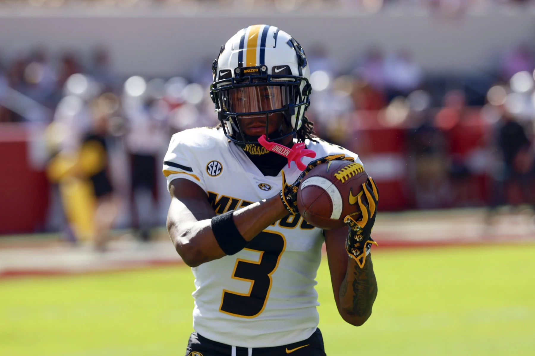 Luther Burden III trägt eine schwarz-weiße Uniform mit der Nummer 3 und demonstriert explosives Potenzial, als er einen Football fängt. Seine Handschuhe weisen rosa Akzente auf. Das verschwommene Spielfeld und die Stadionsitze deuten an diesem sonnigen Tag das Rot im Hintergrund an. Diese Beschreibung wurde mit der FootballR KI automatisch generiert.