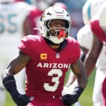 Budda Baker in einer roten Arizona Cardinals-Uniform mit der Nummer 3 auf dem Trikot jubelt auf dem Feld. Er trägt einen weißen Helm mit einem roten Vogel-Logo. Im Hintergrund sind andere Spieler und ein verschwommenes Spielfeld zu sehen, was die Intensität des Augenblicks einfängt. Diese Beschreibung wurde mit der FootballR KI automatisch generiert.