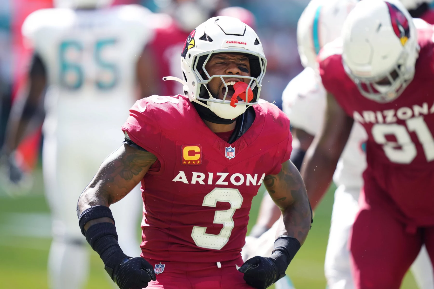 Budda Baker in einer roten Arizona Cardinals-Uniform mit der Nummer 3 auf dem Trikot jubelt auf dem Feld. Er trägt einen weißen Helm mit einem roten Vogel-Logo. Im Hintergrund sind andere Spieler und ein verschwommenes Spielfeld zu sehen, was die Intensität des Augenblicks einfängt. Diese Beschreibung wurde mit der FootballR KI automatisch generiert.