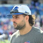 Ein Mann mit Bart und langen Haaren trägt ein graues T-Shirt mit einem Büffel-Logo, das an Matt Milanos Teamgeist erinnert. Er trägt eine weiß-blaue Kappe mit einer Sonnenbrille auf der Krempe, vor einer Stadionkulisse voller verschwommener Zuschauer bei einem spannenden Sportereignis. Diese Beschreibung wurde mit der FootballR KI automatisch generiert.